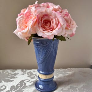 May include: A blue ceramic vase with a textured leaf design and a beige rope accent. The vase holds a bouquet of large, pink roses. The roses have a gradient of pink and white petals.