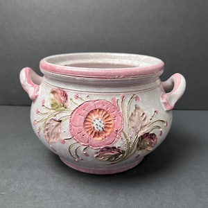 May include: A ceramic pot with two handles, featuring a white base with a raised, hand-painted floral design in shades of pink, orange, and green. The rim and base are accented with a pink border, and the floral motif includes textured petals.