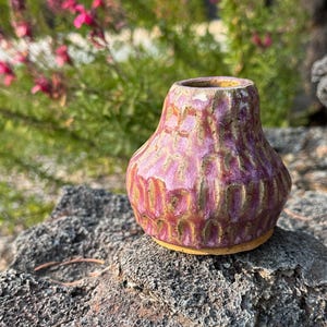 May include: A small, hand-formed ceramic vase with a wide base and narrow neck. The exterior features a textured, dimpled surface finished in a vibrant magenta and purple glaze with exposed tan clay showing through the deep grooves.