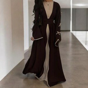 Op de afbeelding: Een lange, donkerbruine jurk met een cr&egrave;mekleurige binnenkant en witte bies. De jurk heeft lange mouwen met strikdetails en een V-hals. De jurk is vloerlang en heeft een vloeiend ontwerp.