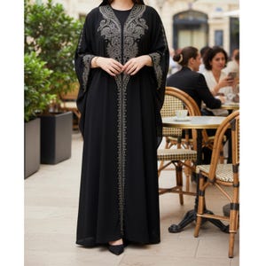 Op de afbeelding: Een zwarte, vloerlange kaftan met ingewikkelde zilveren borduursels langs de halslijn, voorkant en mouwen. Het vloeiende kledingstuk heeft een bescheiden ronde halslijn en wijde mouwen. De kaftan wordt buiten gedragen.