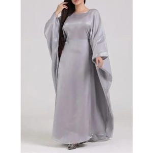 May include: A long, flowing, silver-gray dress with wide, draped sleeves. The dress has a round neckline and a floor-length skirt. The fabric appears to be a shiny, silky material. The overall style is elegant and modest.