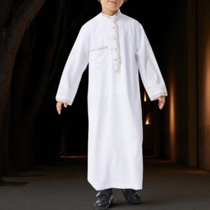 Può includere: Un thobe bianco a maniche lunghe con ricami dorati intorno al colletto, alla tasca e ai polsini. Il thobe è un capo tradizionale, spesso indossato in Medio Oriente e Nord Africa. L'indumento è indossato da una persona.
