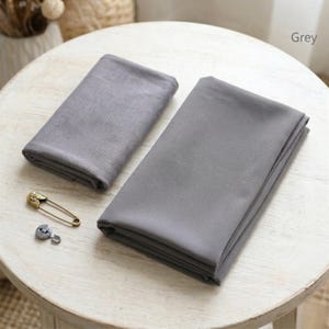 May include: Two folded gray scarves are displayed on a white, round table. A gold safety pin and a small, silver button are next to the scarves. The word "Grey" is in the top right corner.