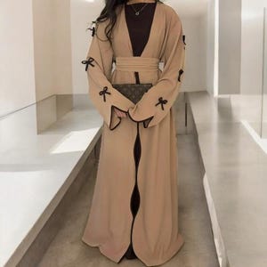 Op de afbeelding: Een lange, vloeiende beige abaya met zwarte strikaccenten op de mouwen. De abaya heeft een bijpassende riem en een donkerbruine onderjurk. Een clutch met patroon wordt vooraan vastgehouden.