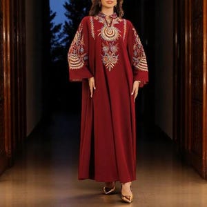 May include: A long, flowing burgundy dress with intricate embroidery in gold, blue, and orange around the neckline and sleeves. The dress features a high collar and wide sleeves, paired with gold-colored heels.