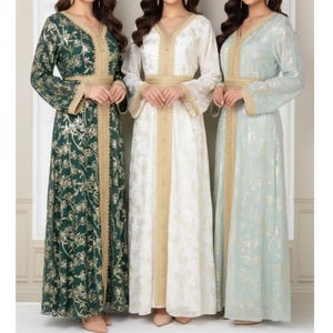 Peut inclure: Trois longues robes fluides, vert émeraude, blanche et bleu pâle. Chaque robe présente un col en V, une garniture dorée et une ceinture dorée assortie. Les robes ont des manches longues et sont en tissu léger à motifs.