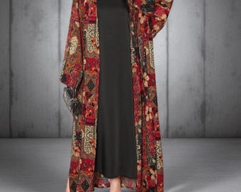 Op de afbeelding: Een lange zwarte jurk wordt gedragen onder een kimono met patroon en open voorkant. De kimono heeft een patchwork-ontwerp met rode, bruine en beige bloemen- en geometrische patronen. De outfit wordt gecompleteerd met zwarte sandalen.