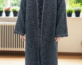 Op de afbeelding: Een lange, open jas in een zwart-witte tweedachtige stof. De jas heeft franjes aan de manchetten en zoom. De jas wordt over een zwarte outfit gedragen. De achtergrond omvat een raam met planten en een radiator.