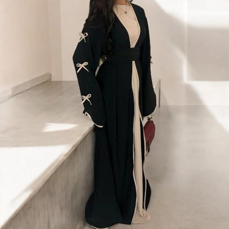 Op de afbeelding: Een zwarte abaya met een cr&egrave;mekleurig interieur en afwerking. De lange mouwen hebben witte strikken en de jurk heeft een vloeiend silhouet. Een bordeauxrode clutch wordt in de hand gehouden.