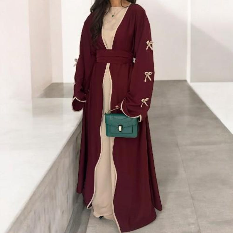 Op de afbeelding: Bordeaux abaya met een beige binnenlaag en bijpassende riem. De mouwen zijn versierd met kleine strikjes. Een kleine, felgroene handtas wordt vooraan vastgehouden.