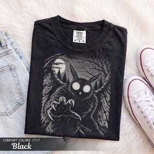 Puede incluir: Una camiseta negra con una ilustración gráfica de un búho con ojos blancos brillantes y garras extendidas. El fondo muestra una escena forestal con luna llena y árboles desnudos. La etiqueta interior dice Comfort Colors M M M.
