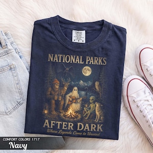 May include: A navy blue t-shirt featuring a graphic print with the text "NATIONAL PARKS AFTER DARK Where Legends Come to Unwind". The illustration depicts a campfire scene with a sasquatch, a ghost playing guitar, an alien, and a wolf.