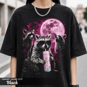 May include: Black t-shirt featuring a graphic of three raccoons under a pink moon. One raccoon wears sunglasses and drinks from a can. The text "Hustle" is visible on the can. The shirt is labeled "Comfort Colors 1717 Black".