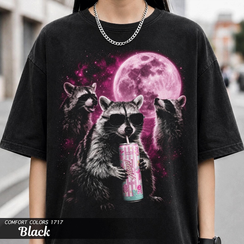 May include: Black t-shirt featuring a graphic of three raccoons under a pink moon. One raccoon wears sunglasses and drinks from a can. The text "Hustle" is visible on the can. The shirt is labeled "Comfort Colors 1717 Black".