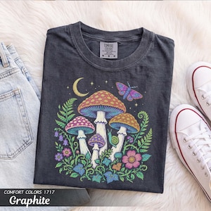 May include: A charcoal gray t-shirt featuring a graphic print of colorful mushrooms, a crescent moon, a butterfly, and a snail surrounded by ferns and flowers. The shirt has a crew neckline and a Comfort Colors brand label on the inside collar.