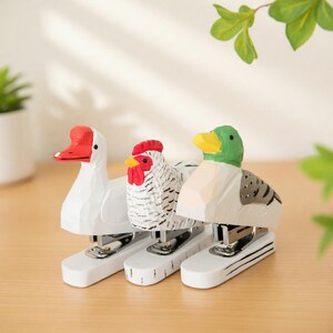 May include: Three whimsical staplers shaped like a duck, chicken, and goose. The duck has a green head and yellow beak, the chicken has a red comb, and the goose has a red beak. Each stapler is white with black accents.
