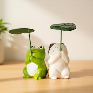 May include: Two decorative figurines: a green frog and a white cat, each holding a green leaf umbrella. The frog has a red mouth and black eyes. The cat has black and red markings and closed eyes. Both are on a wooden surface.