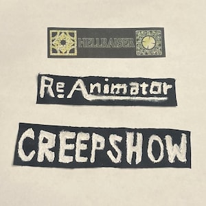 May include: Three black fabric patches with white lettering. The top patch reads "HELLRAISER" with a gold geometric design on each end. The middle patch says "Re-Animator". The bottom patch says "CREEPSHOW".