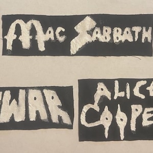 May include: Three rectangular black fabric patches with white embroidered text. The top patch reads Mac Sabbath. The bottom left patch reads GWAR. The bottom right patch reads Alice Cooper. Each patch features raw, unfinished edges and bold lettering.