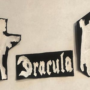 May include: Three black patches with white painted designs. One patch features the word "Dracula" in a gothic font. The other two patches depict silhouettes of Dracula, one in a standing pose and the other in a walking pose.