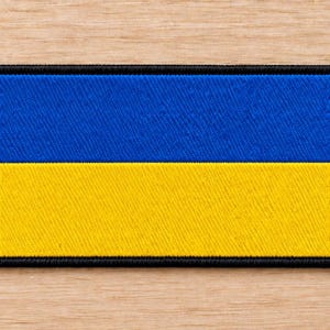 May include: A rectangular embroidered patch featuring the horizontal bicolor design of the Ukrainian flag, with a vibrant blue upper half and a bright yellow lower half, finished with a thick black stitched border around the entire perimeter.