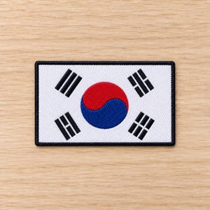 Puede incluir: Un parche rectangular bordado que presenta la bandera de Corea del Sur. El diseño incluye un fondo blanco, un símbolo taegeuk central rojo y azul, y cuatro trigramas negros colocados en las esquinas, todo bordeado por un grueso borde negro.