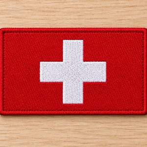 May include: A rectangular embroidered patch featuring a vibrant red background with a centred white cross. The patch has a stitched border and a textured fabric surface, displaying the iconic Swiss flag design in a clean, high-contrast format.