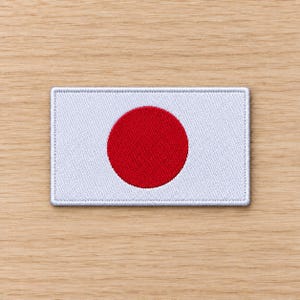 Peut inclure: Un écusson rectangulaire brodé représentant le drapeau national du Japon. Le motif se compose d'un fond blanc uni avec un grand cercle rouge centré. L'écusson présente une bordure cousue et une surface en tissu texturé avec des détails de fils.