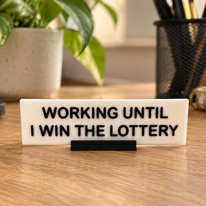 Op de afbeelding: Een rechthoekig wit bureau-bordje met zwarte verhoogde letters met de tekst WORKING UNTIL I WIN THE LOTTERY. Het bordje wordt ondersteund door een kleine zwarte voet en heeft een strak ontwerp voor op een werkplek of bureau.