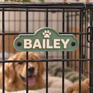 May include: A sage green rectangular name sign with a rounded top edge, featuring the name BAILEY in raised cream-coloured capital letters and a small paw print icon above the text. The sign is attached to a black metal wire crate with two screws.