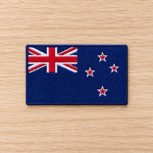 May include: A rectangular embroidered patch featuring the flag of New Zealand. The design consists of a dark blue field with a Union Jack in the upper left corner and four five-pointed red stars with white borders arranged in a Southern Cross pattern.