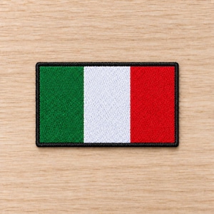 May include: A rectangular embroidered patch featuring the Italian flag design with vertical stripes of green, white, and red. The patch has a thick, solid black stitched border that frames the tricolour fabric, creating a clean and defined edge.