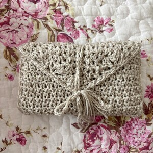 May include: A beige crochet clutch with a flap closure and a tassel tie. The handmade bag features an open weave design and is displayed on a floral patterned fabric. The neutral color and texture of the clutch make it a versatile accessory.