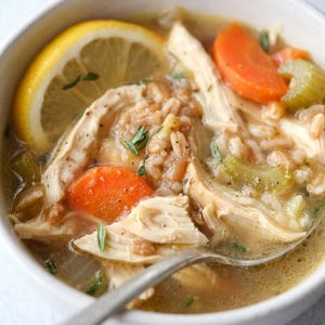 May include: A close-up of chicken soup in a white bowl. The soup contains shredded chicken, orange carrot slices, celery, and barley. A lemon slice and sprigs of herbs garnish the soup. A silver spoon is in the soup.