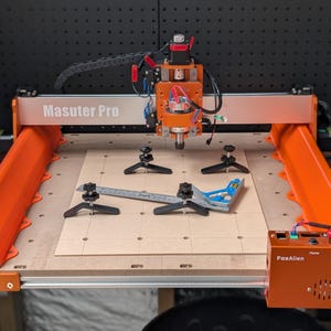 May include: An orange and silver Masuter Pro CNC machine with a wooden base. The machine has a control box labeled FoxAlien and includes a ruler and angle measuring tool. The background is a dark pegboard.