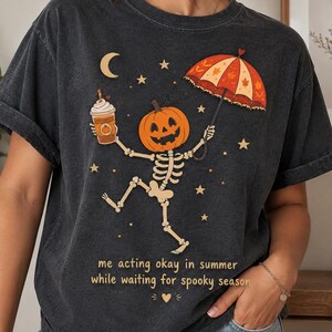 Può includere: Una t-shirt grigio antracite con la grafica di uno scheletro con testa di zucca, che tiene in mano una tazza di caffè e un ombrello arancione a motivi. Il design include stelle, una luna crescente e il testo: me acting okay in summer while waiting for spooky season.