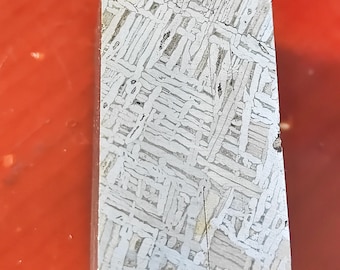 Può includere: Una fetta rettangolare e lucidata di meteorite con un motivo di Widmanstätten. La superficie metallica mostra un complesso disegno a tratteggio incrociato in tonalità argento e grigio. Il taglio rivela l'unica struttura cristallina del materiale extraterrestre.