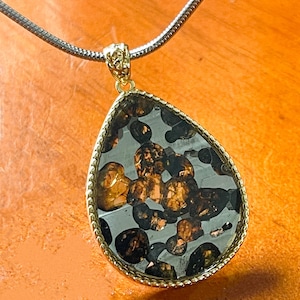 Puede incluir: Un colgante en forma de lágrima con un marco dorado y una cadena plateada. El colgante contiene una mezcla de piedras de color marrón oscuro y dorado sobre un fondo transparente. El colgante mide aproximadamente 3,8 cm de largo.