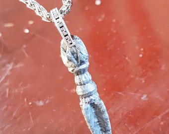 May include: A silver-colored pendant with a textured, elongated design hangs from a silver chain. The pendant has a detailed, carved appearance, with a cylindrical body and a pointed end. The chain is a braided style, and the background is a reddish-brown color.