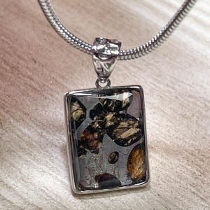 Puede incluir: Colgante rectangular con marco plateado y centro estampado gris oscuro. El patrón incluye formas orgánicas marrones y negras. El colgante cuelga de una cadena plateada.