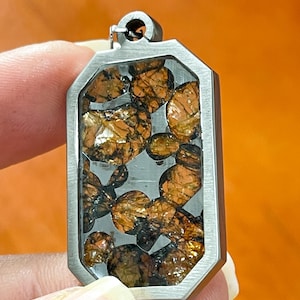 Puede incluir: Un colgante rectangular con un marco plateado. El colgante contiene piedras de color ámbar de forma irregular. Las piedras tienen un aspecto metálico reflectante. El colgante tiene un pequeño lazo en la parte superior para una cadena.