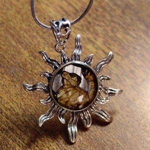 Puede incluir: Un collar con un colgante de sol plateado y una piedra transparente que contiene motas marrones y doradas. El colgante está unido a una cadena plateada.