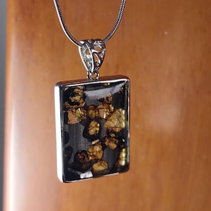 Puede incluir: Un collar de cadena de plata con un colgante rectangular con resina transparente llena de motas doradas.