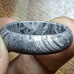 May include: Close-up of a patterned, dark gray ring. The ring has a textured surface with visible lines and patterns. The ring is held between two fingers, showcasing its intricate design and craftsmanship.