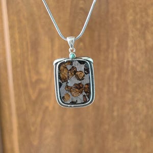 Puede incluir: Un collar de cadena de plata con un colgante rectangular que presenta una piedra con un patrón marrón y plateado. Una pequeña piedra turquesa está engastada en la parte superior del colgante.