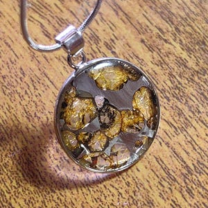 Puede incluir: Un collar con colgante redondo con una cadena plateada. El colgante presenta un mosaico de piezas doradas y plateadas encapsuladas en resina transparente. El colgante mide aproximadamente 2,5 cm de diámetro.