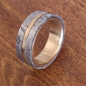 May include: A silver ring with a gold band around the centre. The ring is made of meteorite material and has a unique, textured surface.