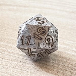 May include: A twenty-sided die featuring a metallic, silver-toned surface with a distinct, layered, wood-grain-like texture. The numbers are deeply engraved into the faces, with the visible digits including 19, 18, 17, 16, 11, and 9.