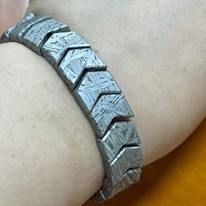 Op de afbeelding: Een armband bestaande uit grijze, pijlvormige metalen segmenten. De armband, gedragen om de pols, heeft een kenmerkende, patroonachtige textuur op elk segment. Een uniek sieraad.
